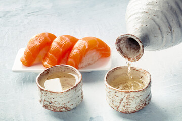 Sake poured into ochoko, traditional cup, with salmon nigiri in the background, traditional Japanese food and drink