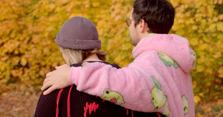 A couple shares a tender moment in an autumn park, surrounded by colorful falling leaves. The guys trendy pink hoodie adds to the relaxed vibe, capturing love and togetherness this season