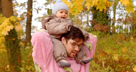 Fototapeta premium In a beautiful autumn forest, an adult carries a cheerful child on their shoulders among vibrant yellow and orange leaves, creating a picturesque scene of joy and warmth