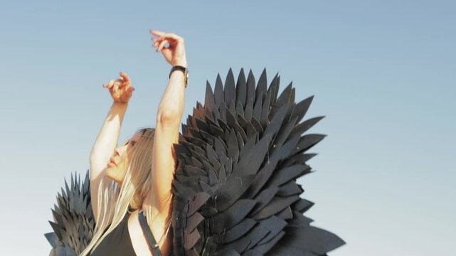 Blonde performer dancing with black angel wings, embodying ethereal movement against bright blue backdrop, expressing celestial grace and dramatic intensity