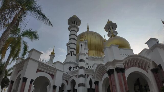Golden-domed mosque with intricate arches, set amidst tall palm trees under a cloudy sky. Ornate towers and lush greenery enhance the majestic architectural scene