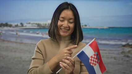 Young woman smiling and pointing to a croatian flag at the seaside, capturing a vibrant beach day with waves and blue sky. - Powered by Adobe