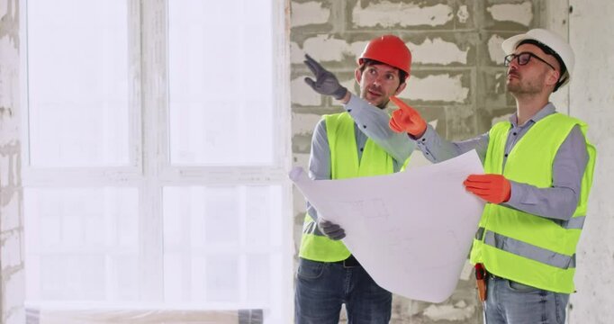 Men experts discuss upcoming work holding floor plan in hands. Builders in helmets and safety vests inspect large apartment and cover work process during conversation
