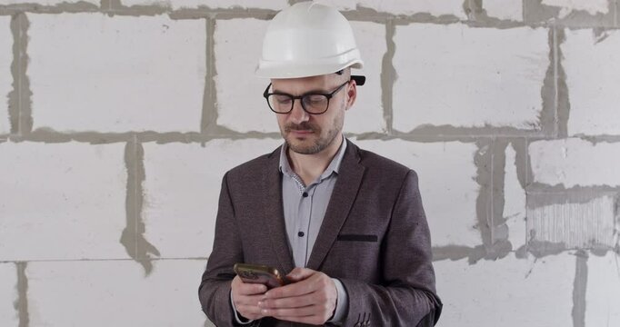Engineer shows frustration with information on smartphone. Specialist in jacket uses modern technologies to solve problem of renovating apartment standing against wall