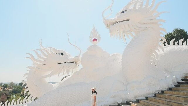 Woman tourist wear stylish white dress sightseeing explores iconic twin white dragons on temple stairs at Wat Huay Pla Kang in Chiang Rai, Thailand under clear blue sky. Famous travel phot spot