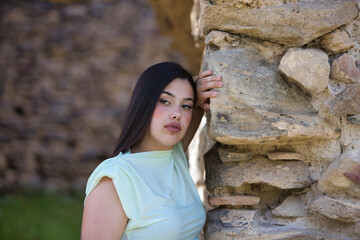 A beautiful Spanish teenager with long dark hair rests her hand on a stone wall and looks sad and...