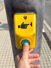 A hand pressing a yellow pedestrian crossing button on a pole. The button features a hand symbol and is located on a sidewalk.
