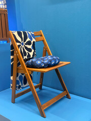 A wooden folding chair with a blue and white floral cushion. The chair is placed against a solid blue background, showcasing a modern design.