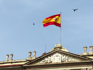 A Spanish flag waves atop a historic building under a clear blue sky. Birds fly in the background,...