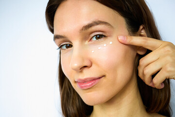 Naklejka premium Beautiful brunette applying eye cream to the area around eyes, beauty portrait on the white background