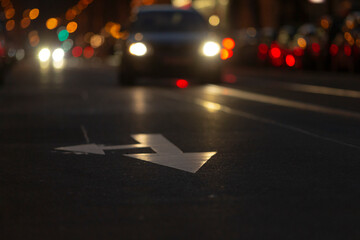 A sleek car approaches on a busy city street at night, marked by a left turn arrow on the asphalt,...