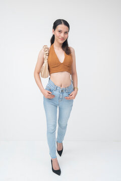 Fototapeta Confident young Filipino woman walking with a stylish outfit, wearing a brown crop top and blue jeans, carrying a bag, isolated on a white background