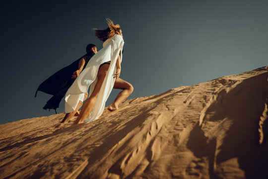 Stylish Couple Climbing Sand Dune in Desert Wind - Powered by Adobe