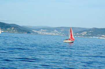 A vibrant red sailboat glides across calm blue waters near a coastal city, framed by rolling hills...