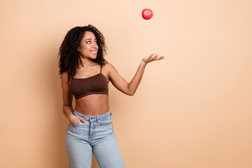 Young woman tossing an apple in the air, smiling confidently against a beige background, promoting...