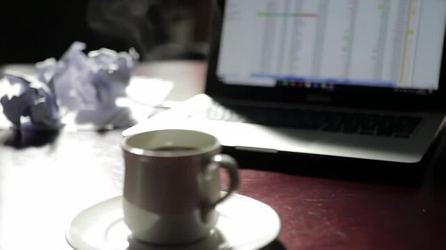 Coffee cup on saucer beside laptop and crumpled papers on table  