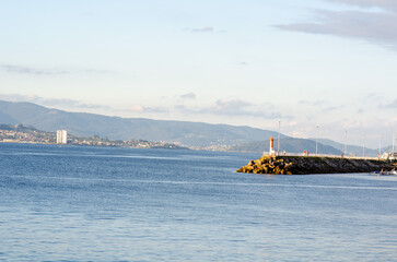 Tranquil scene of a coastal pier with a red lighthouse, calm sea, distant city buildings and green...