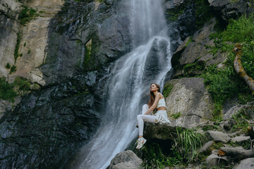 Woman sitting near waterfall in white activewear with relaxed and peaceful expression surrounded by...