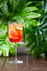 A tall glass of refreshing cocktail with ice cubes, orange slices, and a cucumber slice, set against a vibrant green backdrop of tropical leaves.