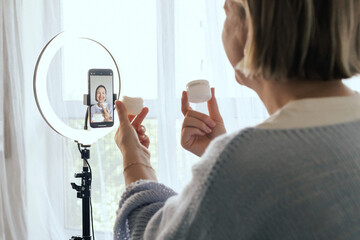 Middle-aged woman beauty blogger filming skincare product unboxing at home with ring light. User-generated content UGC for online store promotion. Vlogger live streaming. Part of a series