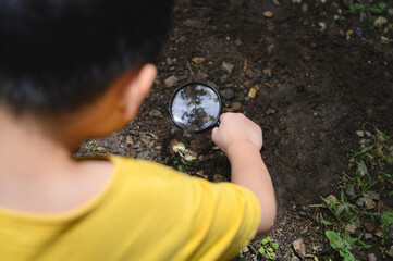 Young boy wearing yellow t-shirt observing ground with magnifying glass in natural outdoor setting,...