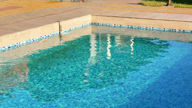 A close-up view of the sparkling blue water of an outdoor swimming pool, showing the mosaic tiles at the bottom and the light-colored pool coping.