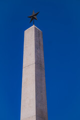 Reggio Emilia, Italy, 01.04.2025.Tall stone obelisk monument with metal star tip against deep blue sky, minimal composition