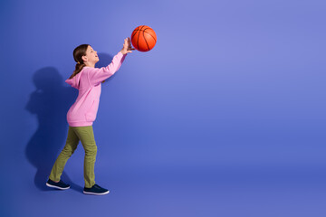Fototapeta premium Young schoolgirl joyfully playing with a basketball against a vibrant purple background, wearing casual pink hoodie and green pants.