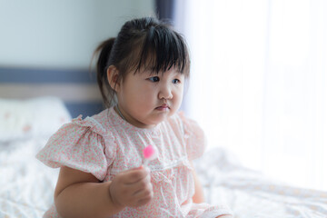 Fototapeta premium A cute little girl is happily eating a lollipop,Sweet tooth Asian girl happy holding big lollipop sitting on bed at home