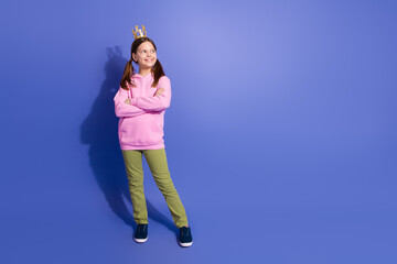 Cheerful preteen girl wearing a pink hoodie and crown standing on purple background with a...