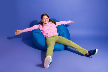 Obraz premium Adorable happy schoolgirl sitting on a comfortable blue bean bag against a vibrant purple background