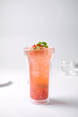 A refreshing pink iced drink garnished with mint and fruit pieces in a clear double-walled glass on a white surface with a blurred background.
