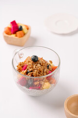 A clear glass cup with chia pudding topped with granola and fresh fruits, with a small bowl of mixed fruit in the background on a white surface.
