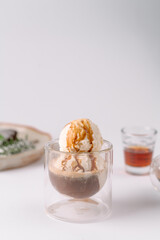 A scoop of vanilla ice cream topped with caramel sauce, served in a double-walled glass, with a neutral background and blurred objects in the distance.