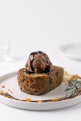 A decadent slice of chocolate bread topped with a scoop of chocolate ice cream and drizzled with chocolate sauce, served on a white plate with decorative crumbs and a garnish.