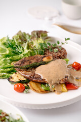 Grilled steak with creamy sauce, served with fresh greens, roasted vegetables, and quinoa on a white plate.