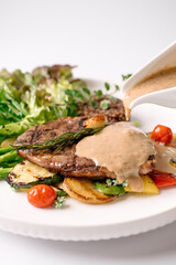 A gourmet steak topped with creamy sauce, served with grilled vegetables and fresh salad on a white plate.