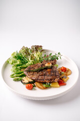 Grilled steak served with roasted vegetables, cherry tomatoes, and fresh greens on a white plate.