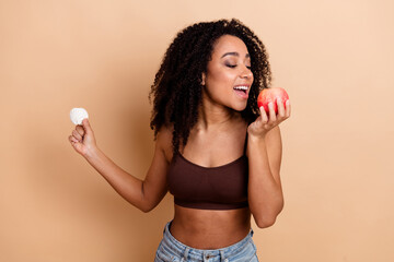 Smiling woman holding apple and candy in hands on beige background showcasing healthy lifestyle and...