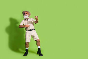 Elderly man in beige safari-style outfit having fun on a green backdrop, displaying wanderlust and adventure with his playful pose