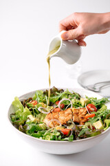 A fresh, colorful salad with grilled chicken is being drizzled with a green dressing from a small pitcher.