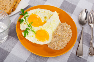 Homemade fresh breakfast - sunny-side up eggs and side dish, oatmeal porridge. Empty glass, fork, knife, bran bread. Table is covered with checkered tablecloth.
