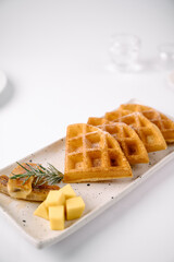 Four golden waffle quarters dusted with powdered sugar, served on a rectangular plate with cheese cubes and a sprig of rosemary.