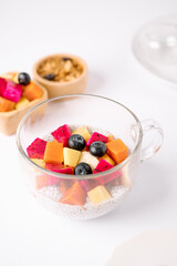 Transparent cup with chia pudding topped with colorful fruit cubes including dragon fruit, papaya, banana, and blueberries on a white surface.