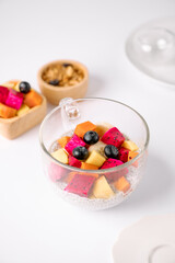 A clear cup filled with a colorful mix of fresh fruits including dragon fruit, blueberries, and papaya, with a small bowl of granola in the background on a white surface.