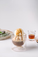 A glass cup containing a scoop of vanilla ice cream with caramel drizzle over a coffee ice sphere, with a small glass of caramel syrup in the background.