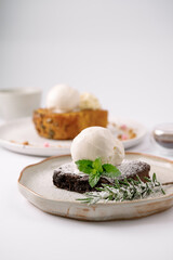 A scoop of vanilla ice cream atop a chocolate brownie garnished with mint and powdered sugar on a ceramic plate, with a blurred dessert and cup in the background.