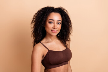 Attractive young woman with natural curls in casual brown top, looking confident against beige...