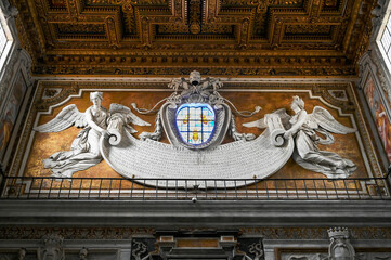 Baroque wall relief inside a Roman church with Latin inscription held by two angels, stained glass...