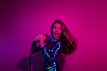 Young woman in glossy blue dress holding microphone, singing with joy against a vibrant pink neon background, festive night out.